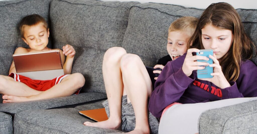 Niños usando tablet y móvil en el sofá, ejemplo de uso excesivo de pantallas en la infancia.